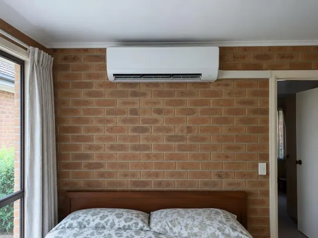 Wall-mounted split system above a bed in a brick bedroom with curtains in Salisbury Downs